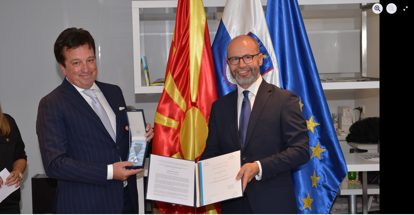 Veleposlanik in odlikovanec stojita pred zastavami obeh držav in proti objektivu držita odlikovanje in plaketo.