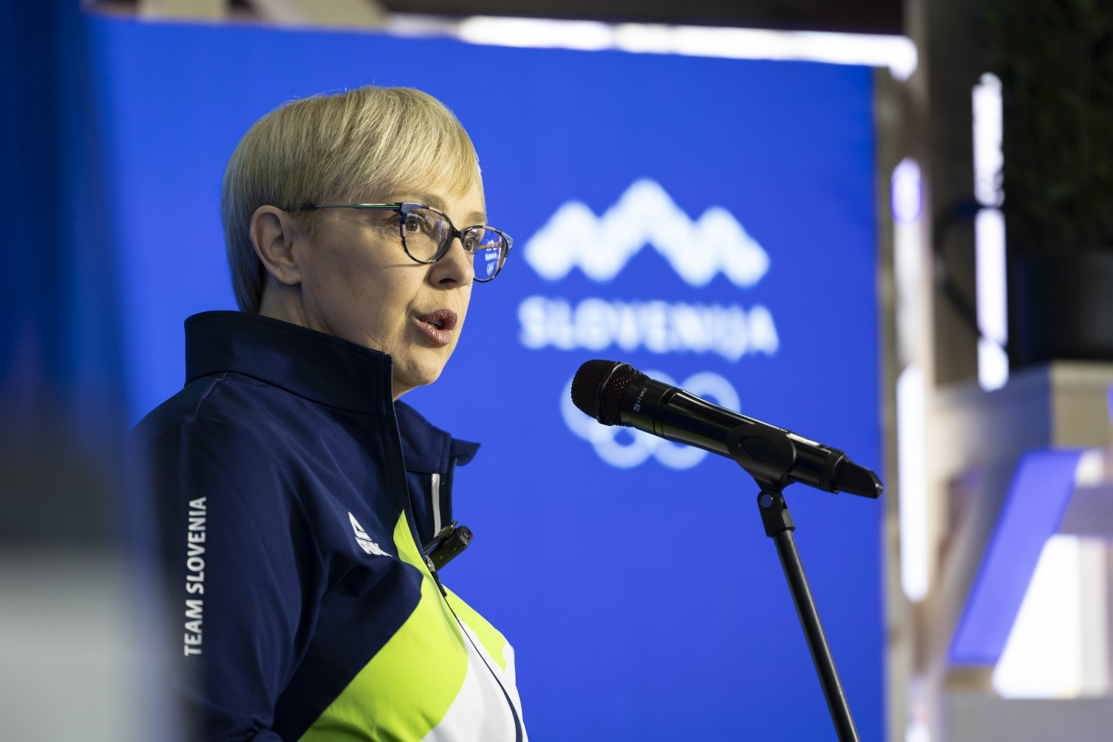 Predsednica stoji pred mikrofonom, v ozadju je napis Slovenija in olimpijski krogi.