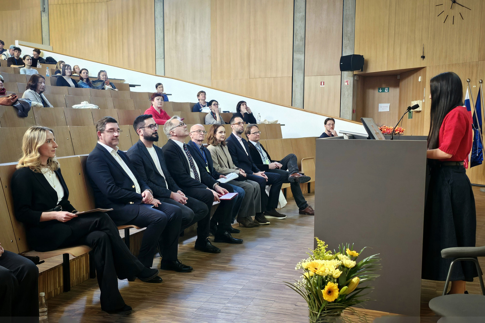 Soprog in drugi udeleženci simpozija sedijo v prvi vrsti v dvorani in poslušajo nagovor.