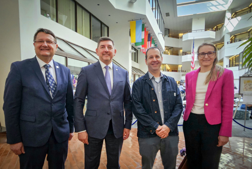 Skupinska fotografija štirih oseb v svetlem atriju z zastavami v ozadju.