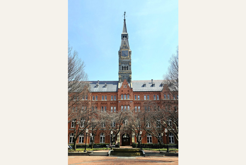 Zgodovinska rdečeopečnata stavba z visokim stolpom in uro, obdana z drevjem.