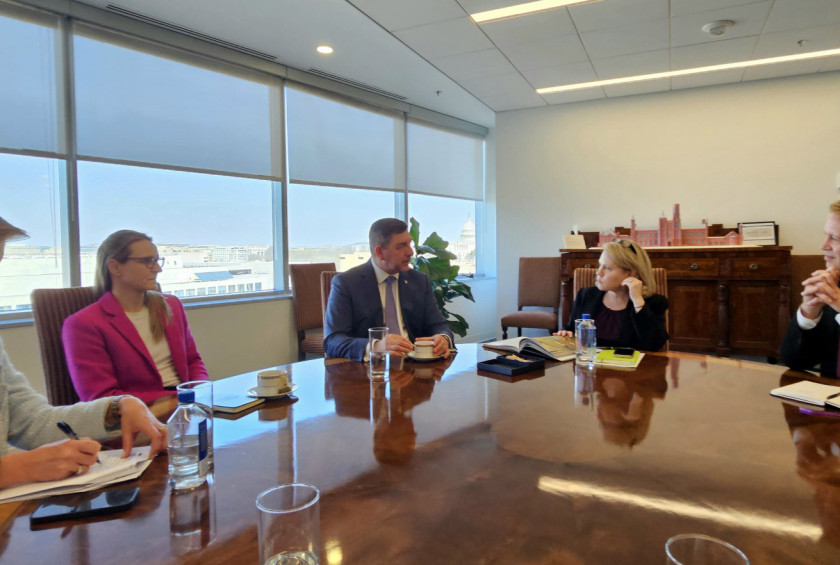 Skupina ljudi sedi za mizo na sestanku, skozi okno se vidi Kapitol v Washingtonu.