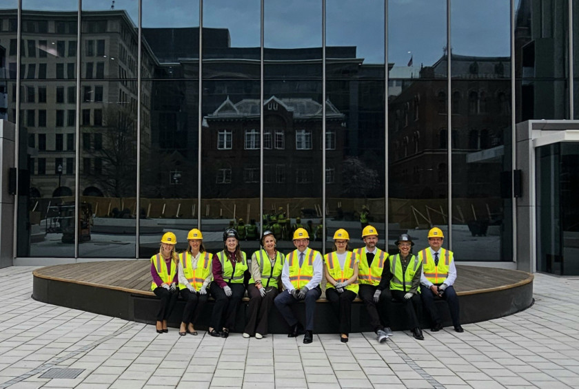 Skupina ljudi v zaščitni opremi sedi pred moderno stavbo s stekleno fasado.