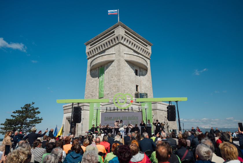 Slovesnost ob stolpu z odrom in občinstvom na prostem, slovenska zastava na vrhu.