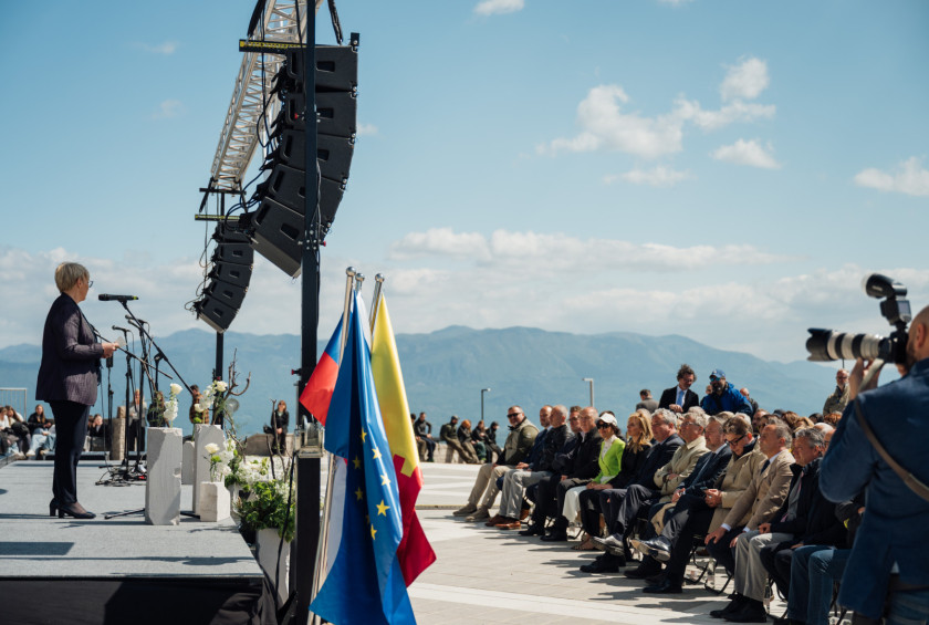Pogled s strani na predsednico, kiima govor na odru in občinstvo, v ozadju gore in zastave.