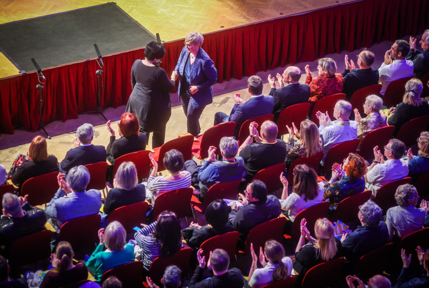Predsednica se rokuje z direktorico JSKD, ljudje, ki sedijo ploskajo.