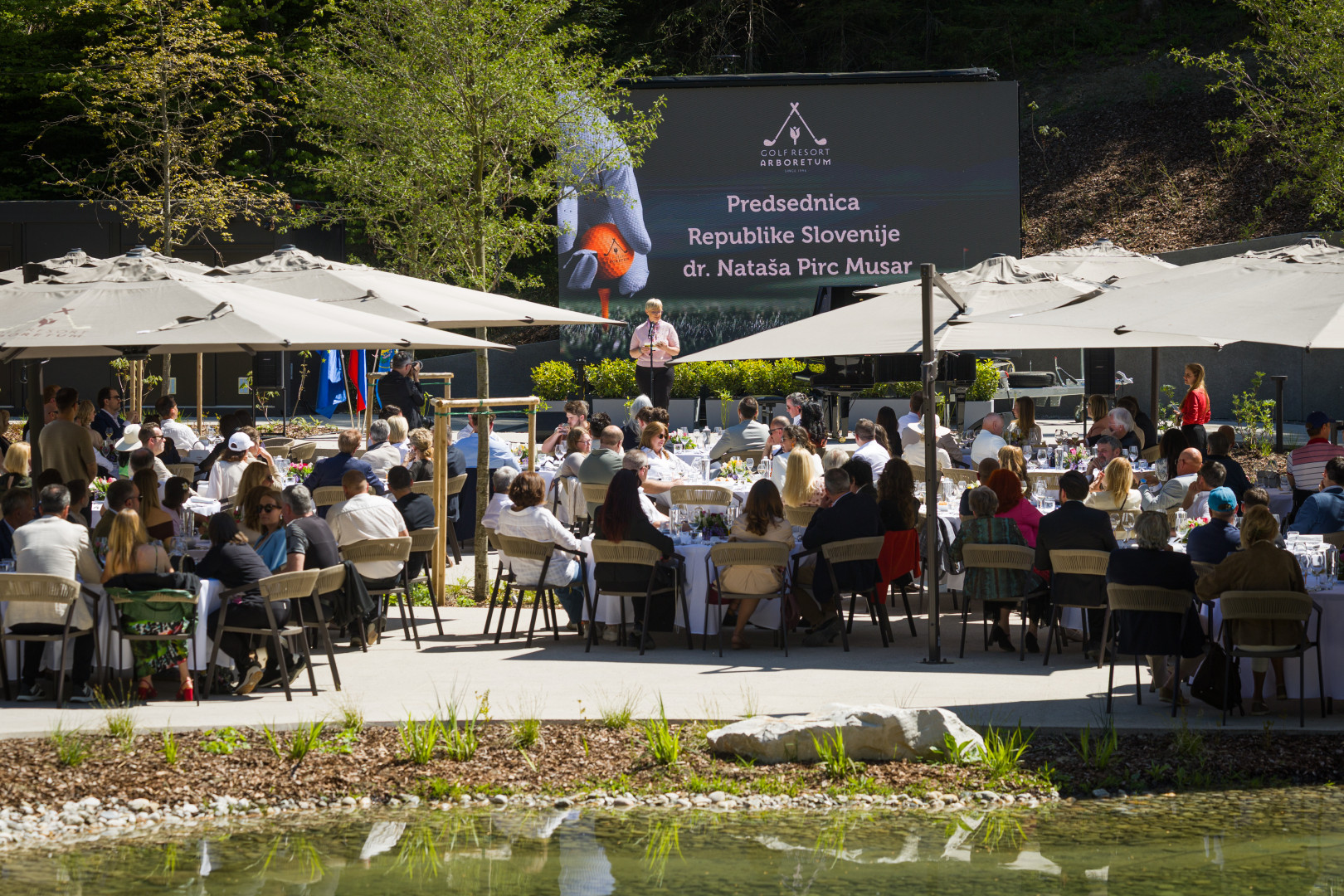 Pogled na zbrane na odprtju novega golf igrišča, predsednica v ozadju stoji na odru.