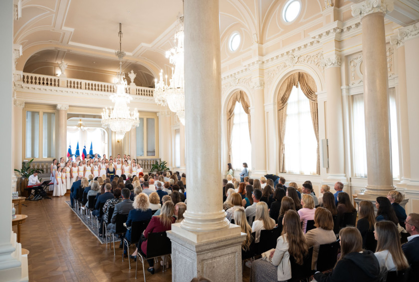 Pogled na Veliko dvorano v kateri sedijo udeležnci in udeleženke sprejema in spremljajo nastop pevskega zbora.
