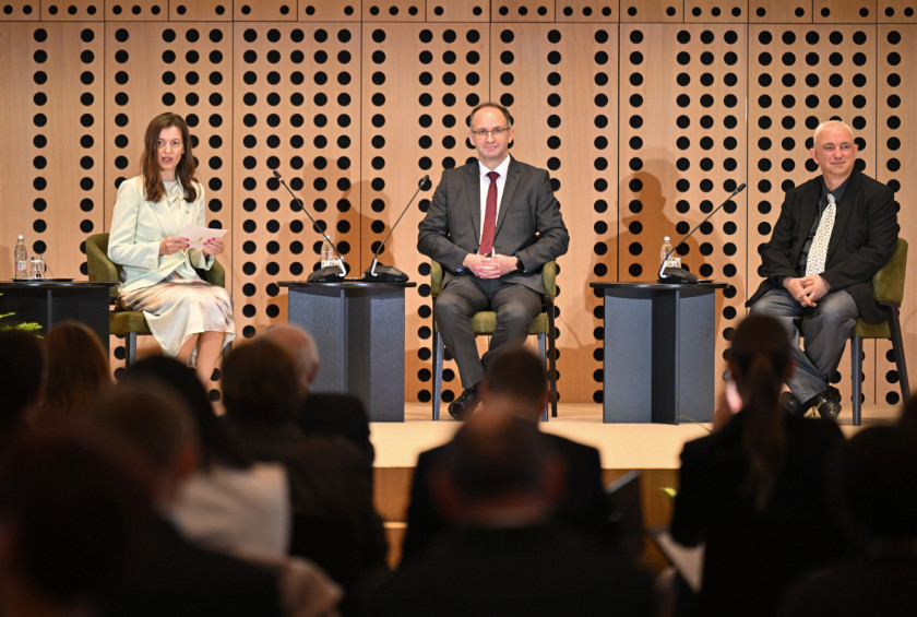 Trije panelisti na forumu sedijo na odru in opazujejo občinstvo.