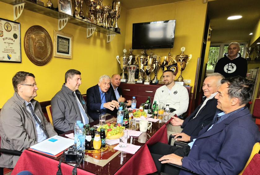 Udeleženci srečanja sedijo za manjšo mizo, na policah okoli njih so številni pokali.