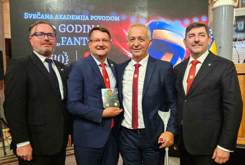 Štirje udeleženci sprejema v temnih oblekah, med njimi je soprog, stojijo pred ozadjem na katerem je napis Svečana akademija.