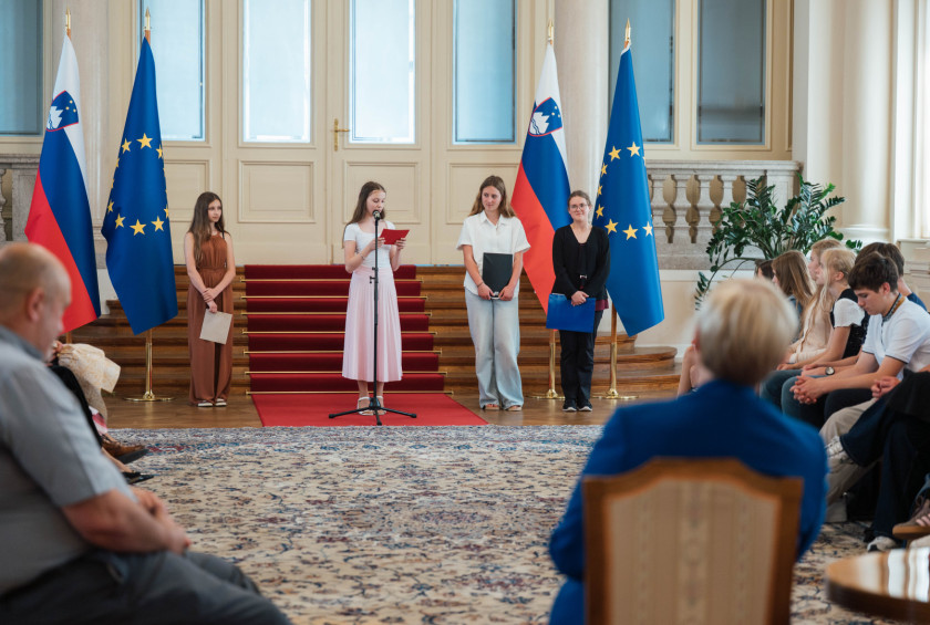 Dekle govori pred mikrofonom, predsednica nasproti nje posluša ob drugih obiskovalcih.