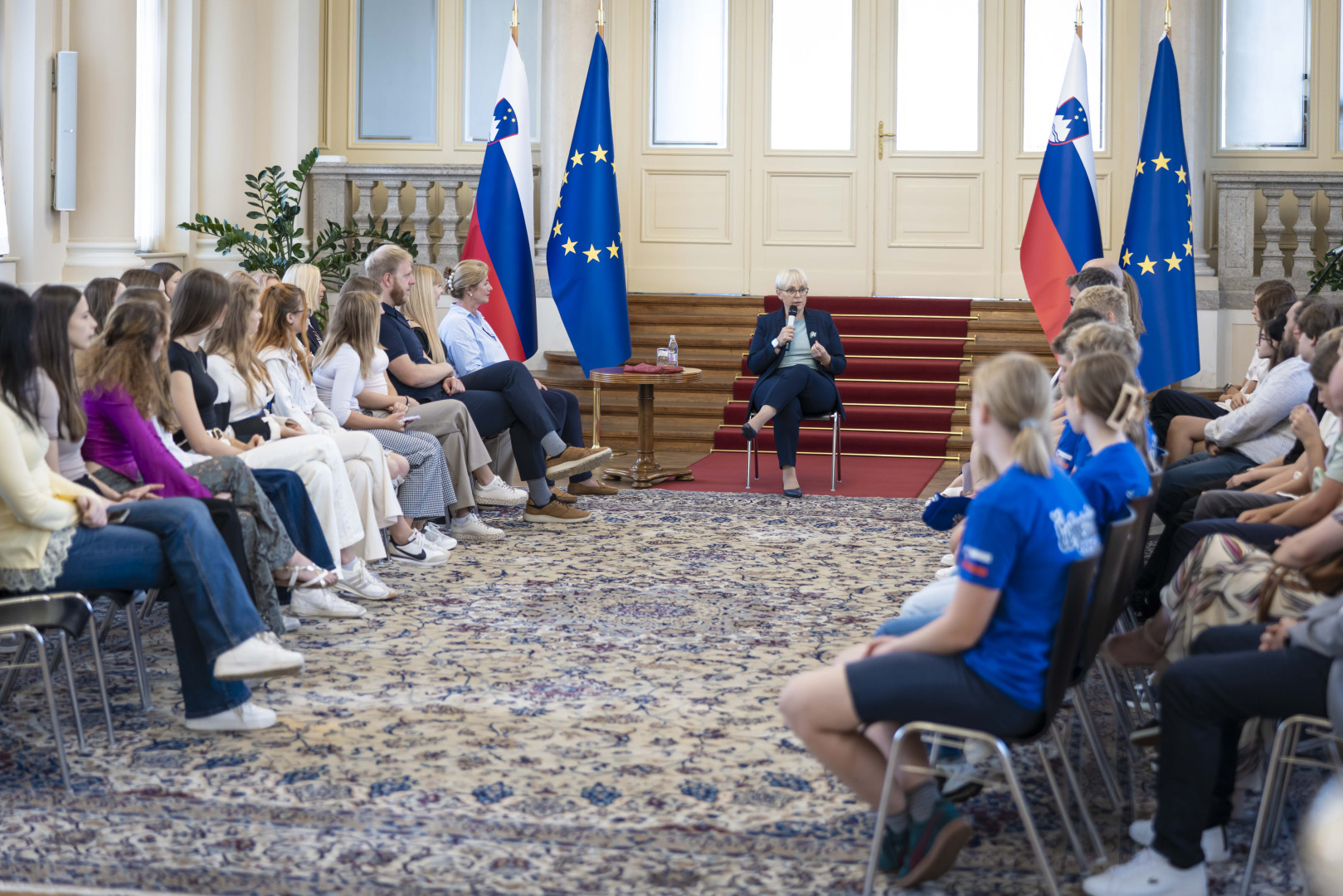 Dijaki in učenci sedijo v polkrogu in spremljajo nagovor predsednice.