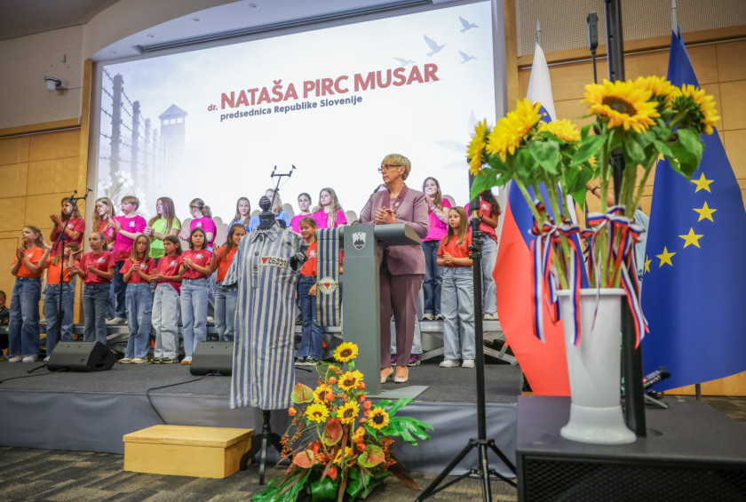 Predsednica na odru nagovarja zbrane na dogodku, v ozadju je otroški pevski zbor.