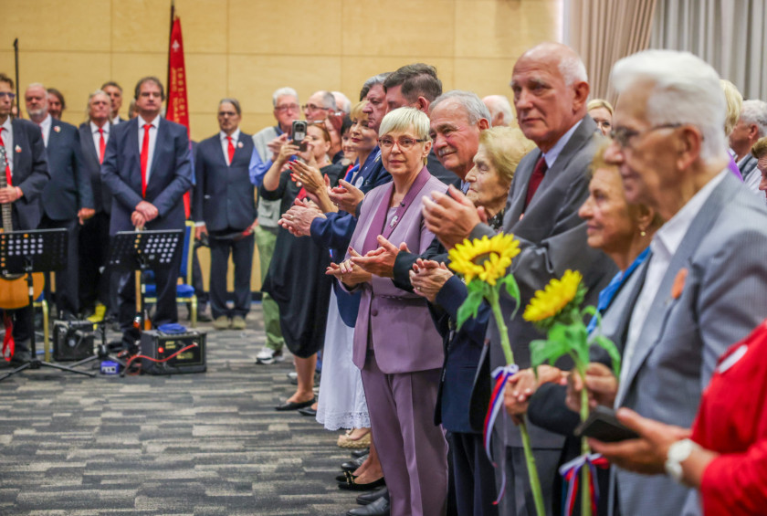 Predsednica in drugi udeleženci dogodka stojijo v prvi vrsti in ploskajo.