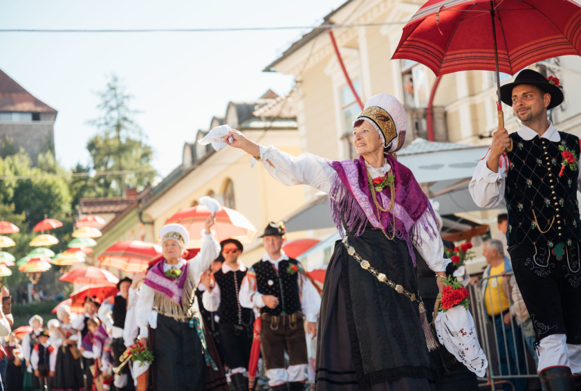 Udeleženci v narodnih nošah sodelujejo v slavnostni povorki skozi staro mestno jedro Kamnika ter nosijo tradicionalne okrasne dežnike.