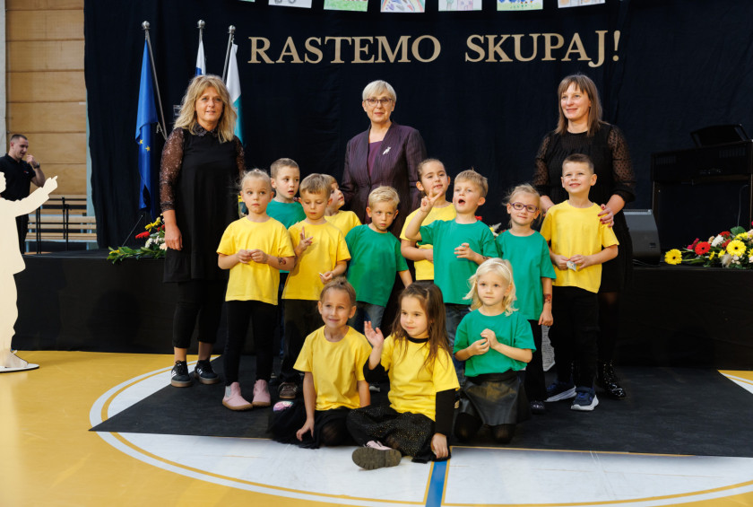 Predsednica stoji na odru s skupino otrok in dvema vzgojiteljicama v zelenih in rumenih majicah.